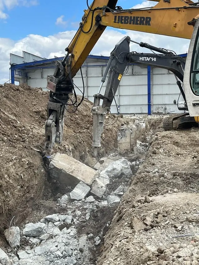 Liebherr kotró osztályozó-törőfejjel és Hitachi kotró hidraulikus bontókalapáccsal vasbeton alapgerendát bont árokban, acélcsarnok előtt
