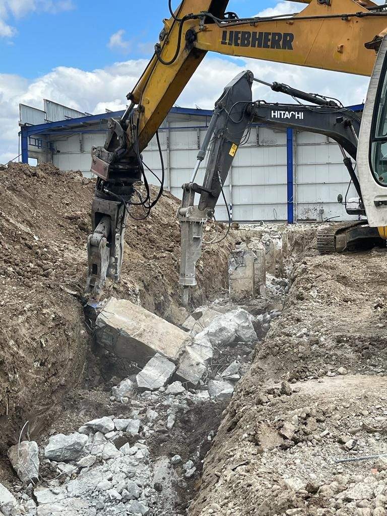 Liebherr kotró osztályozó-törőfejjel és Hitachi kotró hidraulikus bontókalapáccsal vasbeton alapgerendát bont árokban, acélcsarnok előtt