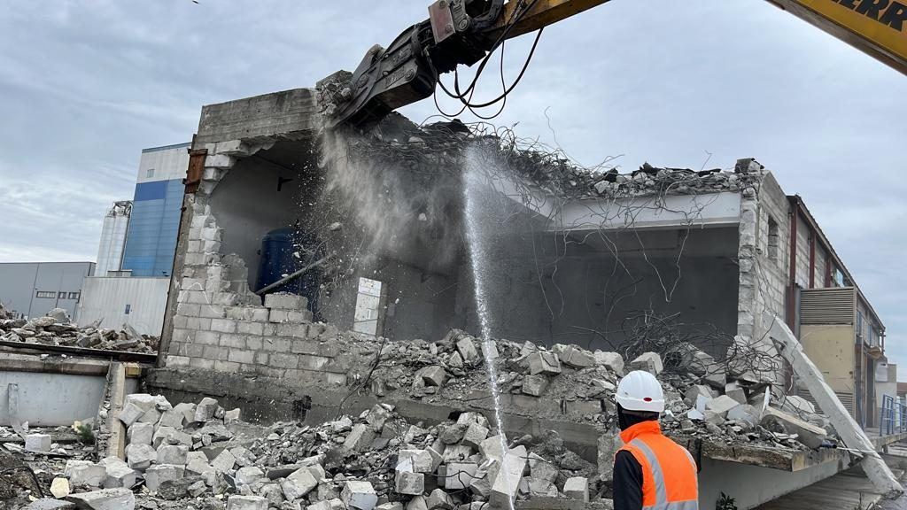 Betonblokk falazatú ipari épület bontása kotróra szerelt bontóollóval, vízpermetes porkötéssel, mellette védősisakos műszaki ellenőr